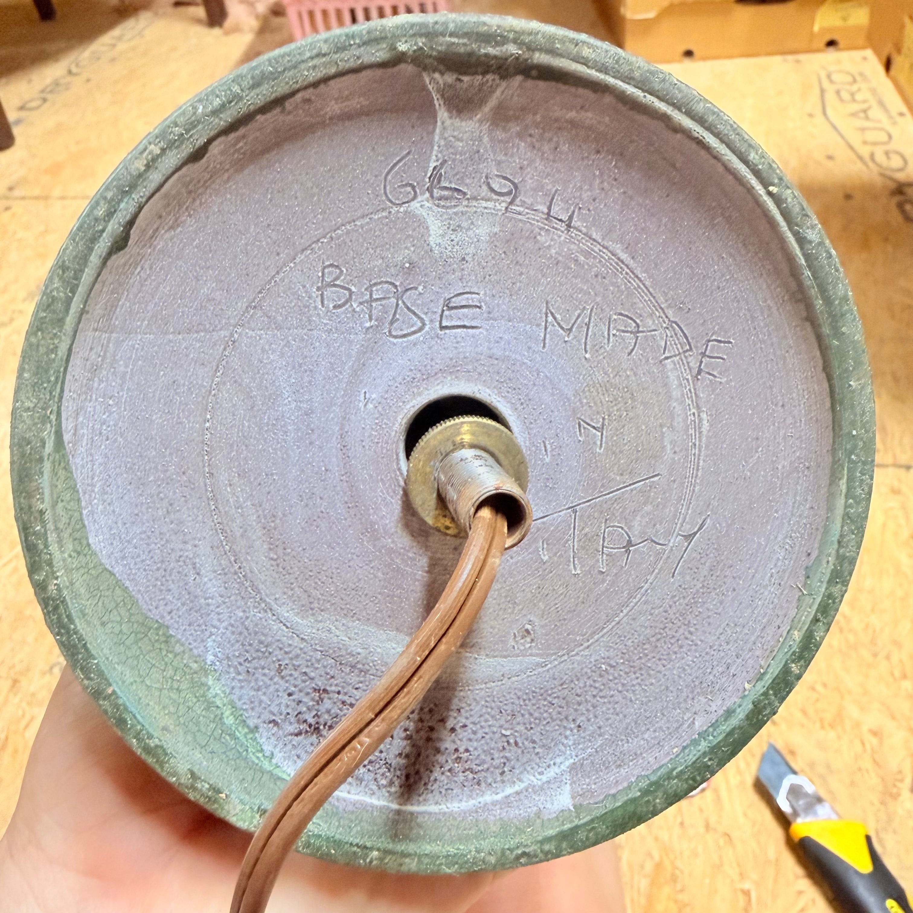 A close-up of a handmade ceramic lamp base with a green glazed rim, a central brass socket with a brown wire attached, and handwritten markings on the back. A hand is holding the object in a workshop setting.