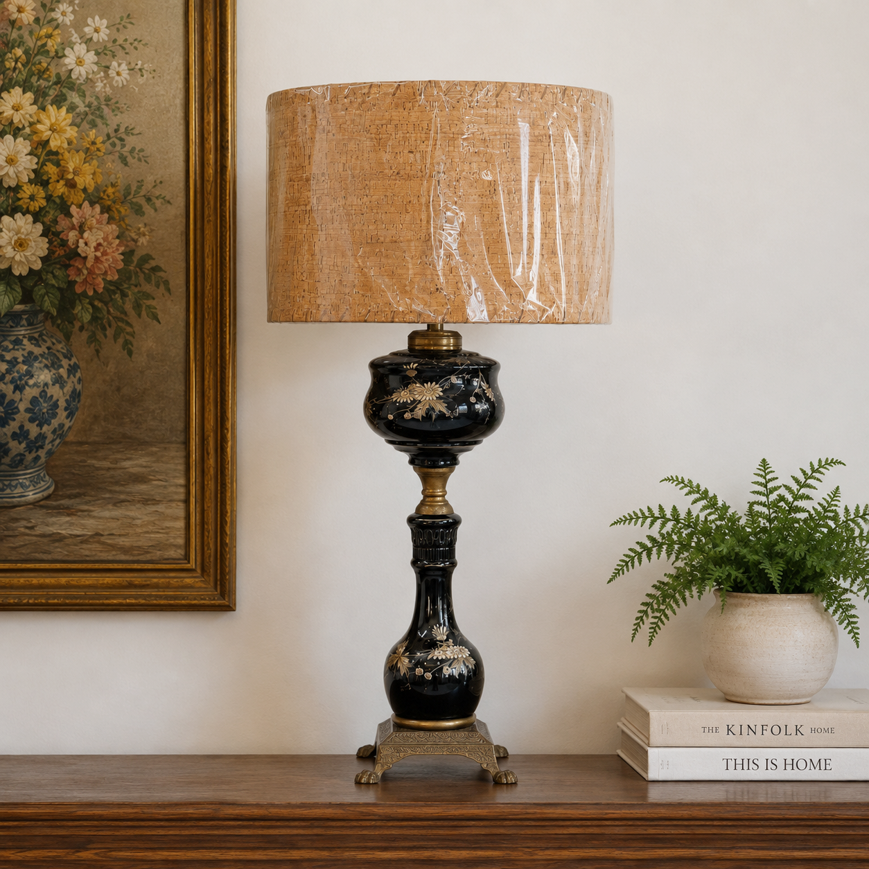 A decorative table lamp with a black and gold painted ceramic base and a beige linen drum shade wrapped in protective plastic, styled on a wooden console table next to a potted fern and a framed floral painting.