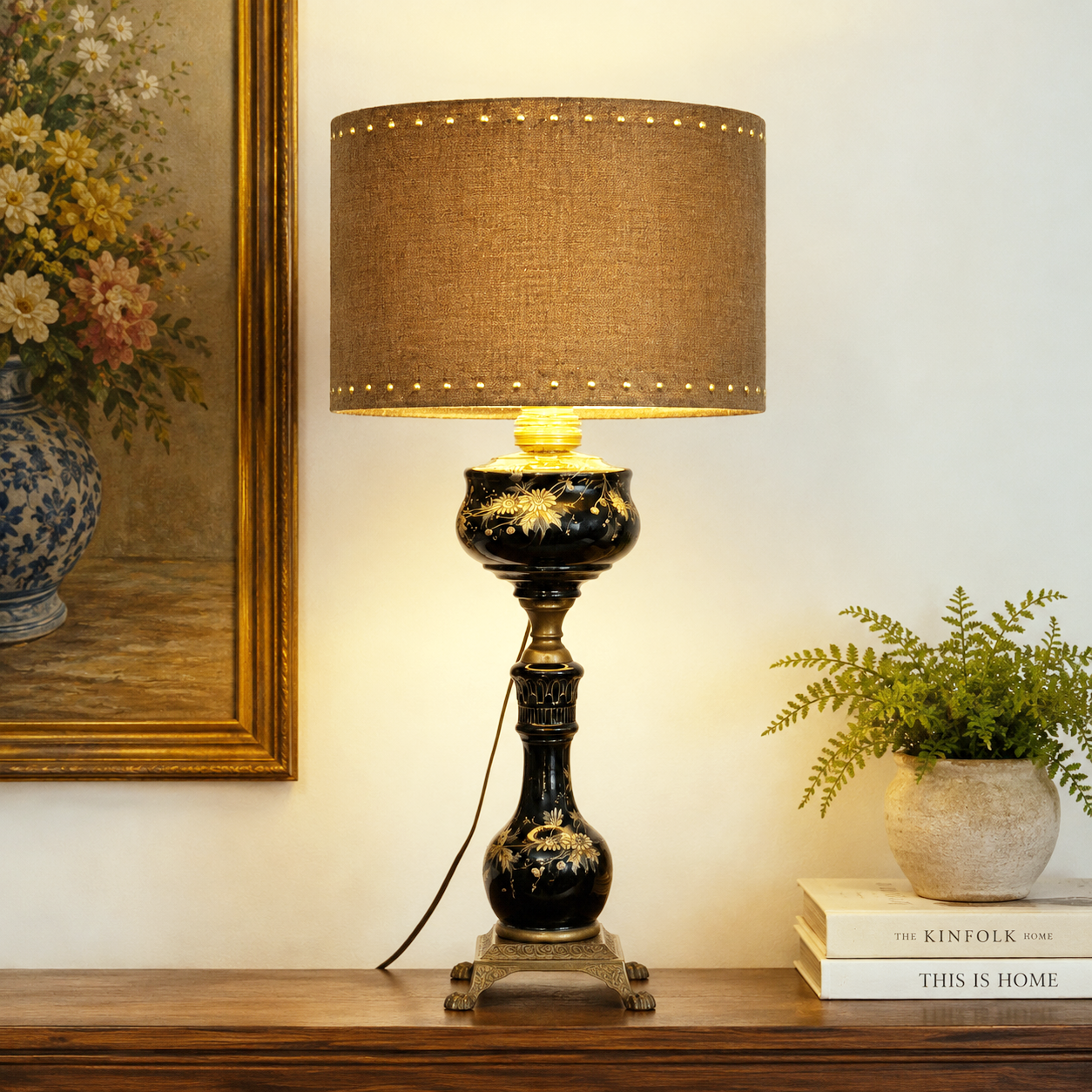 A decorative table lamp with a brown cork drum shade, a black and gold ornate ceramic base, and brass nailhead trim at the top, shown lit on a wooden console against a cream wall with a framed painting and a small potted fern nearby.