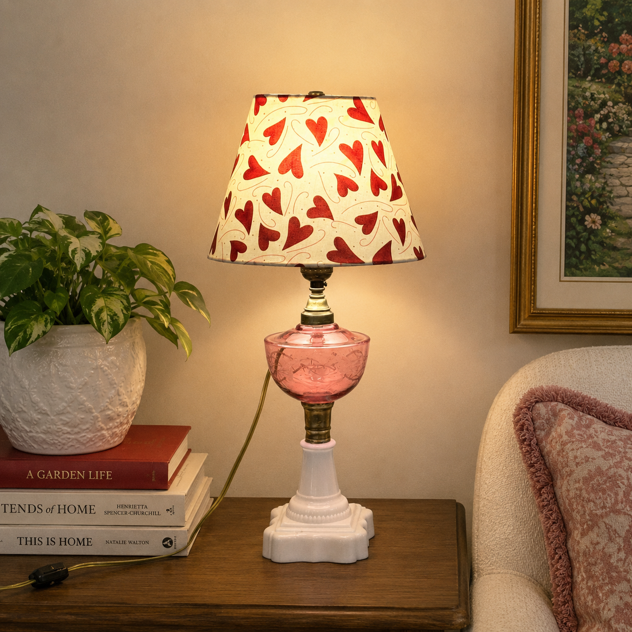 A lit table lamp with a white drum shade covered in red hearts, a pink glass bowl-shaped middle section, and a white pedestal base, placed on a wooden side table in a cozy room setting.