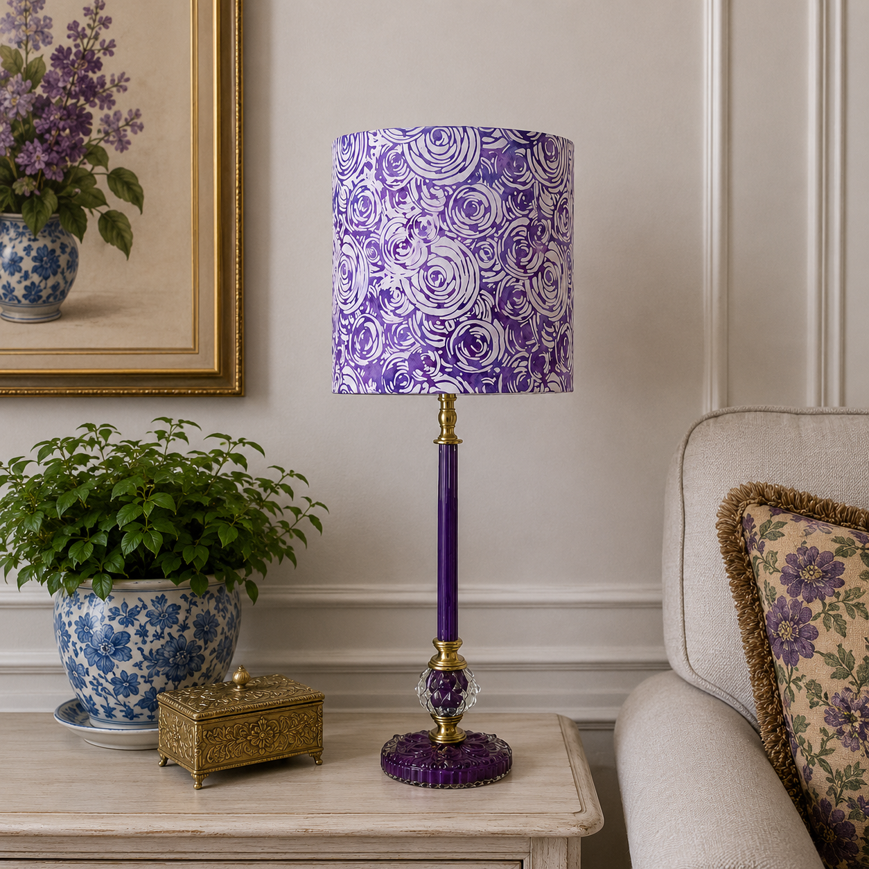 A purple and white floral patterned table lamp with a purple stem-shaped base and gold accents, styled on a light wood side table in a cozy living room setting next to a sofa and a potted plant.