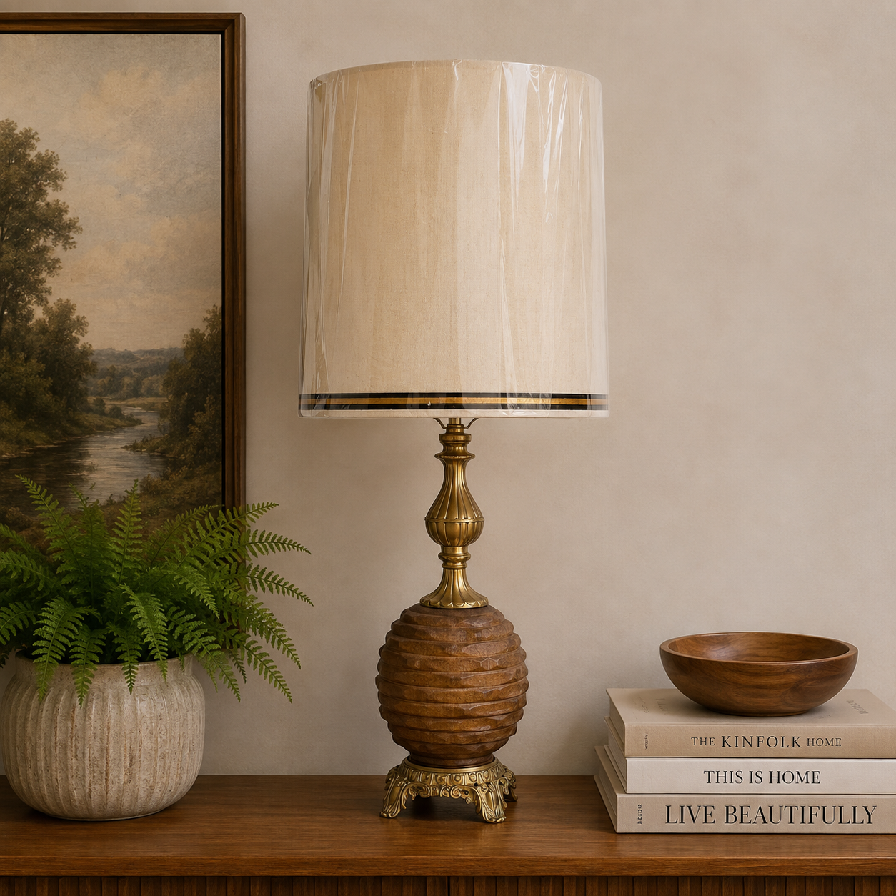 A table lamp with a cream fabric drum shade, a carved wooden ribbed base, and an antique brass finial and pedestal foot, styled on a wooden shelf next to a potted fern and books.