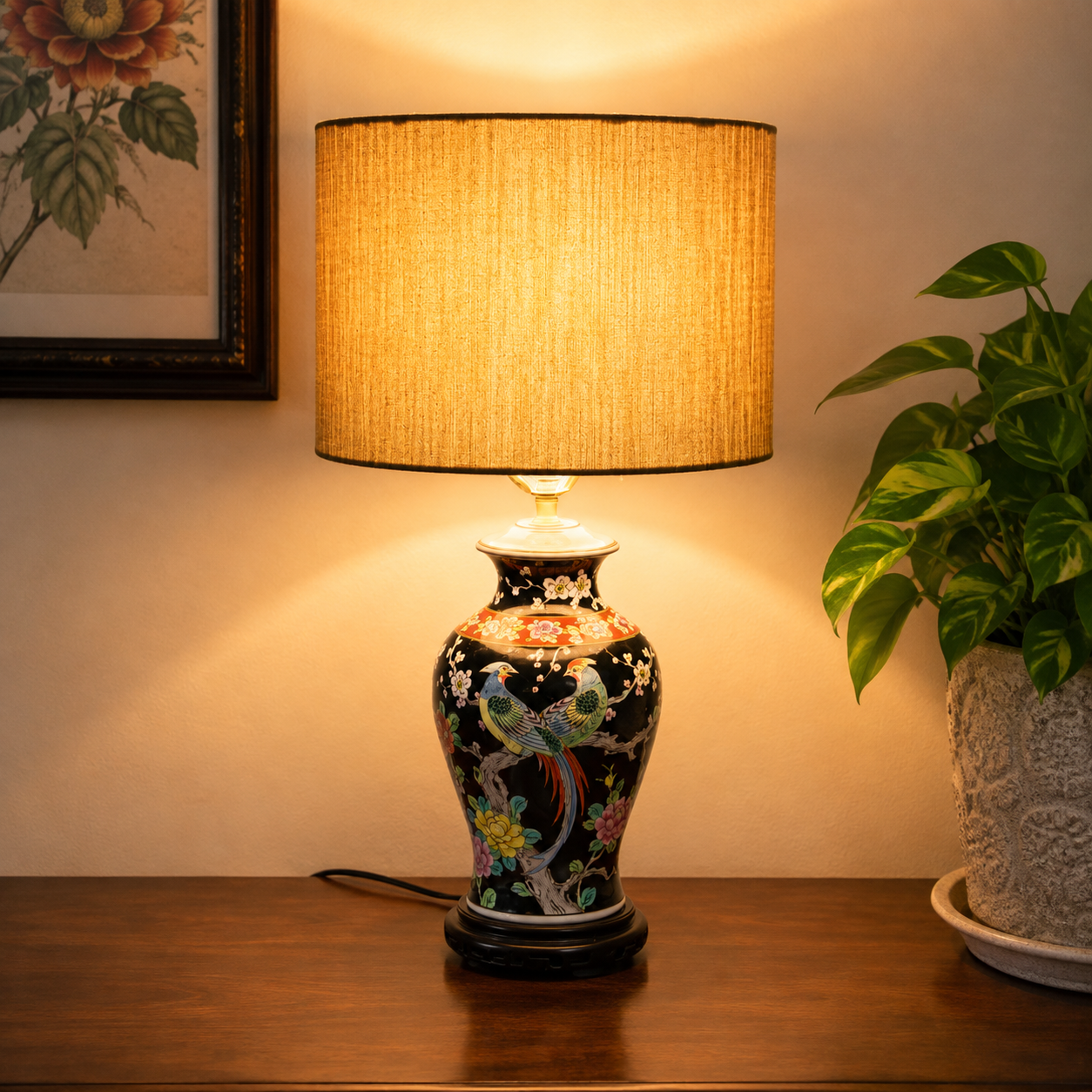 A decorative table lamp with a black porcelain-style base featuring colorful bird and floral motifs, topped with a beige textured drum shade, shown lit on a wooden surface against a warm wall.