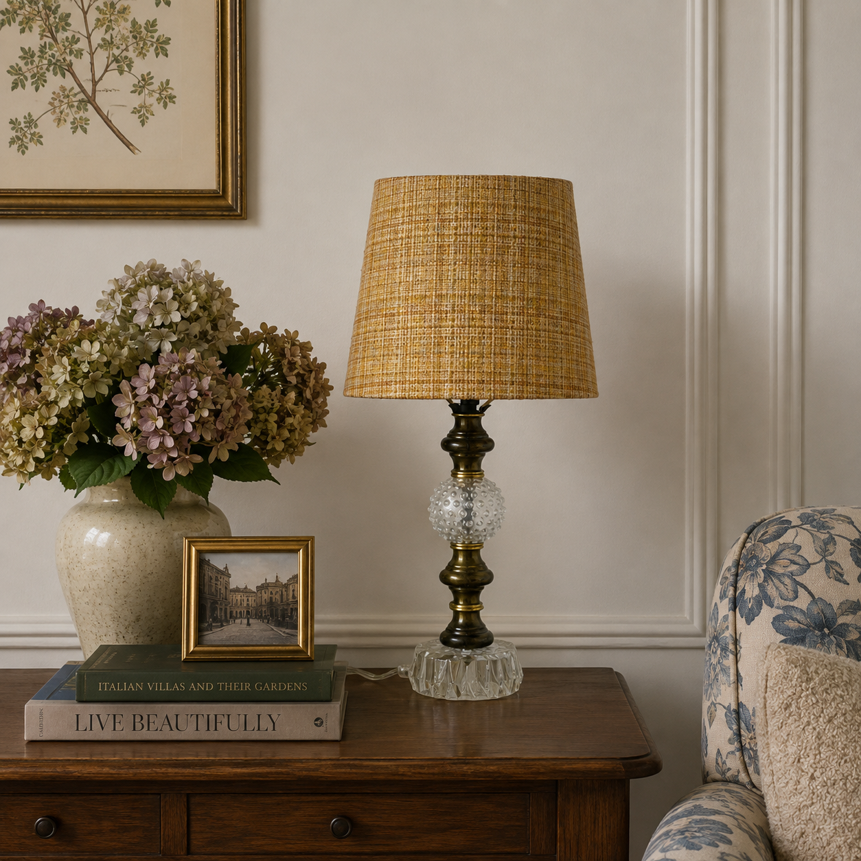A table lamp with a woven straw-colored shade and a clear faceted glass base sits on a wooden side table in a styled room setting with a vase of flowers, books, and a floral armchair.