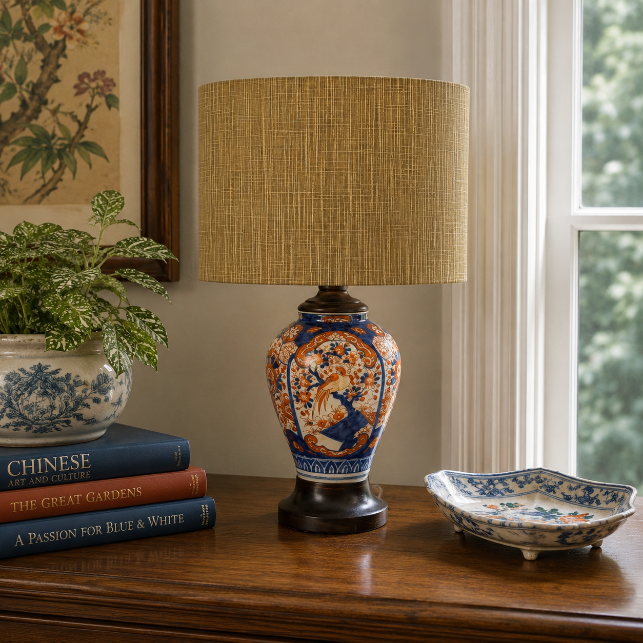 A decorative table lamp with a textured beige linen drum shade and an ornate ceramic base featuring blue, orange, and white floral patterns, styled on a wooden console table next to books and a bowl.