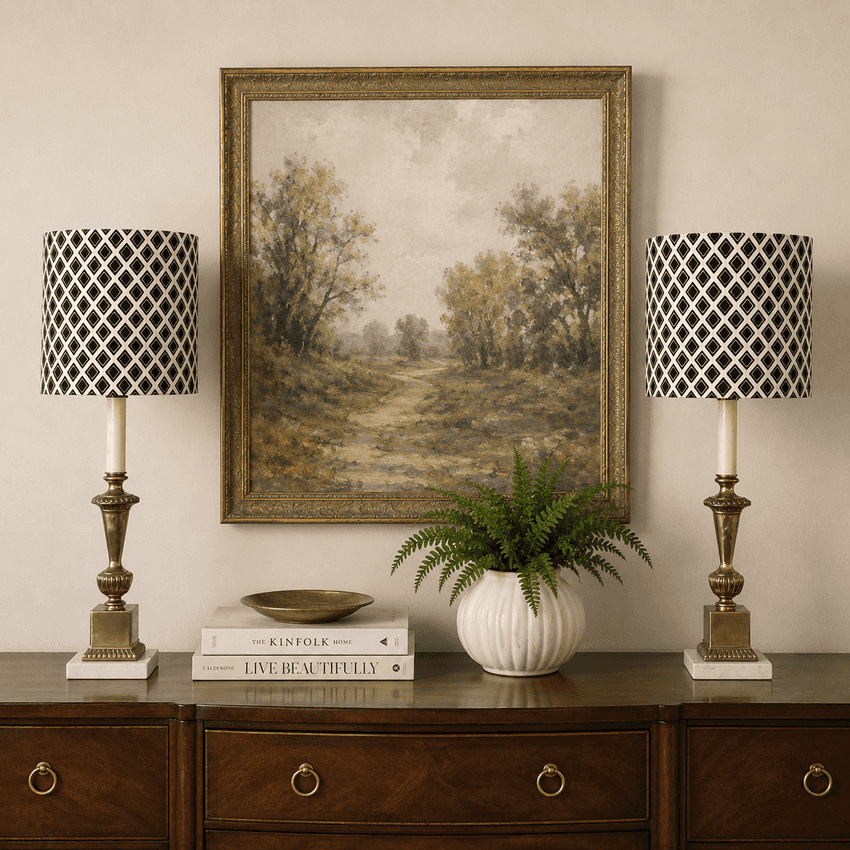 A framed landscape painting hangs on a beige wall above a dark wood console table, flanked by two matching table lamps with black-and-white patterned drum shades and gold-toned bases. A white vase with a green fern and stacked books sit below the artwork.