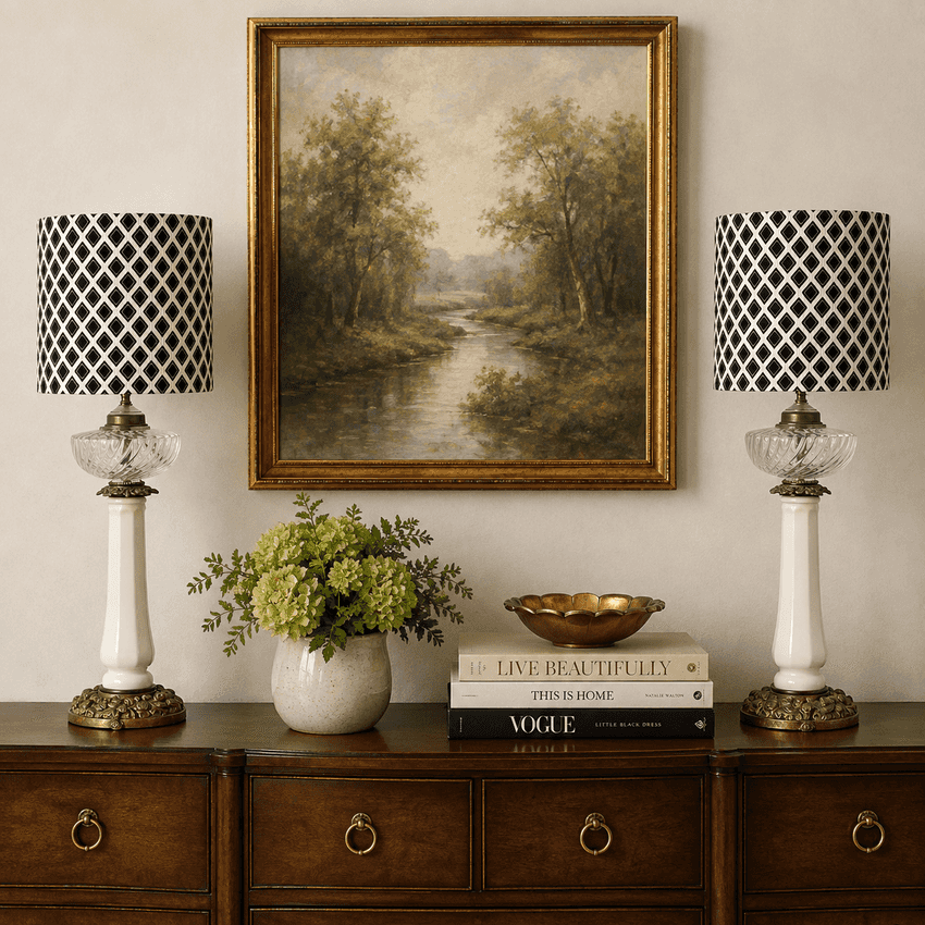 A framed landscape painting of a river flowing through a wooded valley, displayed on a beige wall above a dark wood console table. Two matching table lamps with black-and-white patterned drum shades, white columns, and gold bases sit on either side of the painting.
