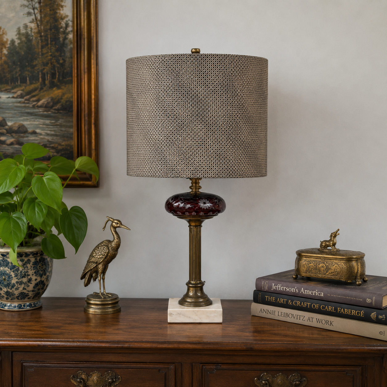 A table lamp with a textured taupe-gray woven drum shade, a dark reddish-brown glass orb base, and a brass-toned pedestal on a white marble base, styled on a wooden console table with a plant, decorative bird figurine, and books.