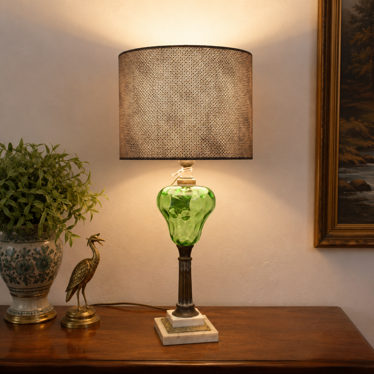 A table lamp with a textured brown drum shade, a green glass bulb, and a dark metal stem on a marble base, styled on a wooden console table next to a potted plant and a framed picture.
