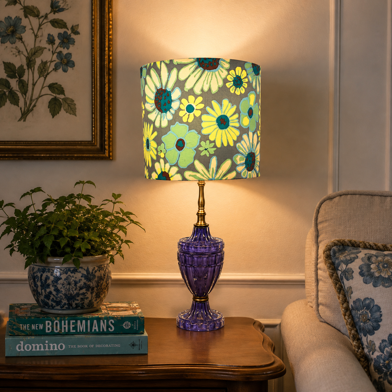 A table lamp with a floral-patterned drum shade in yellow, green, and blue on a gray background, paired with a purple glass base, set on a wooden side table in a cozy living room.