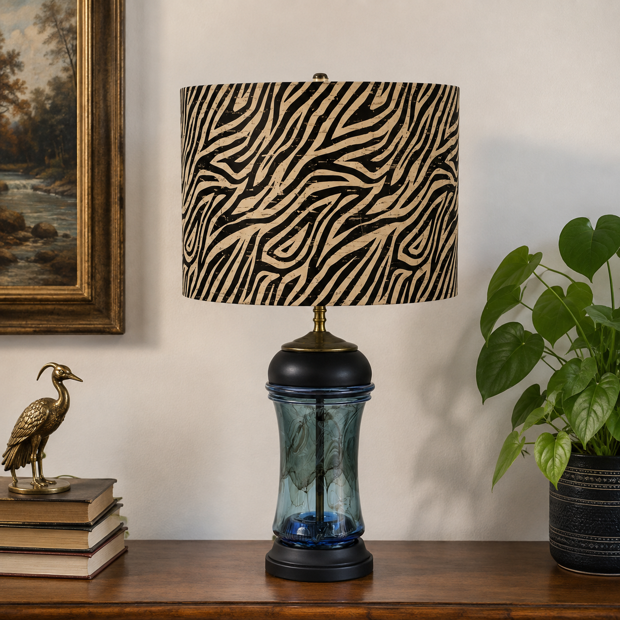 A table lamp with a black and beige zebra-print drum shade, a dark blue glass bulb, and a black base, displayed on a wooden shelf next to a potted plant and a brass bird figurine.