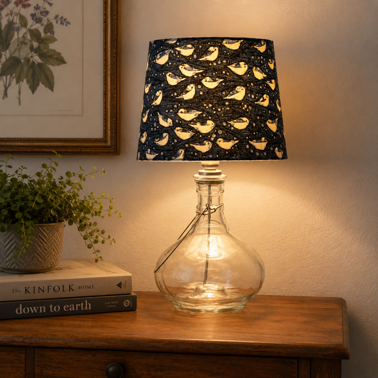 A table lamp with a clear glass bottle base and a dark navy fabric shade printed with a repeating pattern of yellow birds and small stars, lit to cast a warm glow on the wall beside a wooden cabinet.