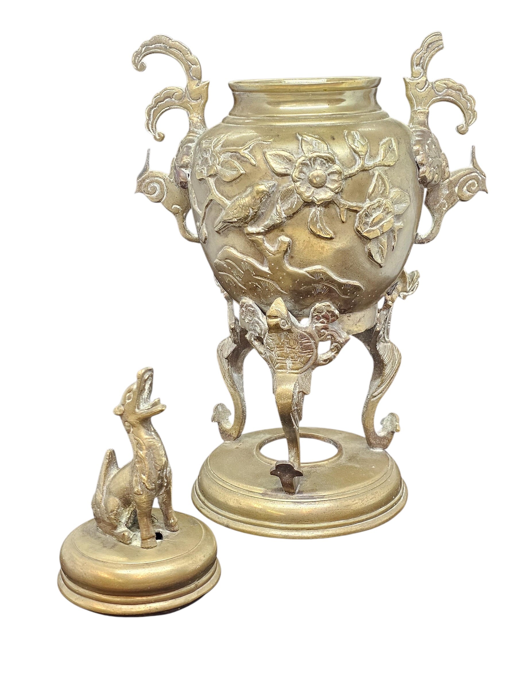 An ornate antique-style brass or bronze urn with sculpted floral and animal motifs, three curved legs, decorative side handles, and a matching small dog-shaped lid or censer placed in front, shown on a plain white background.