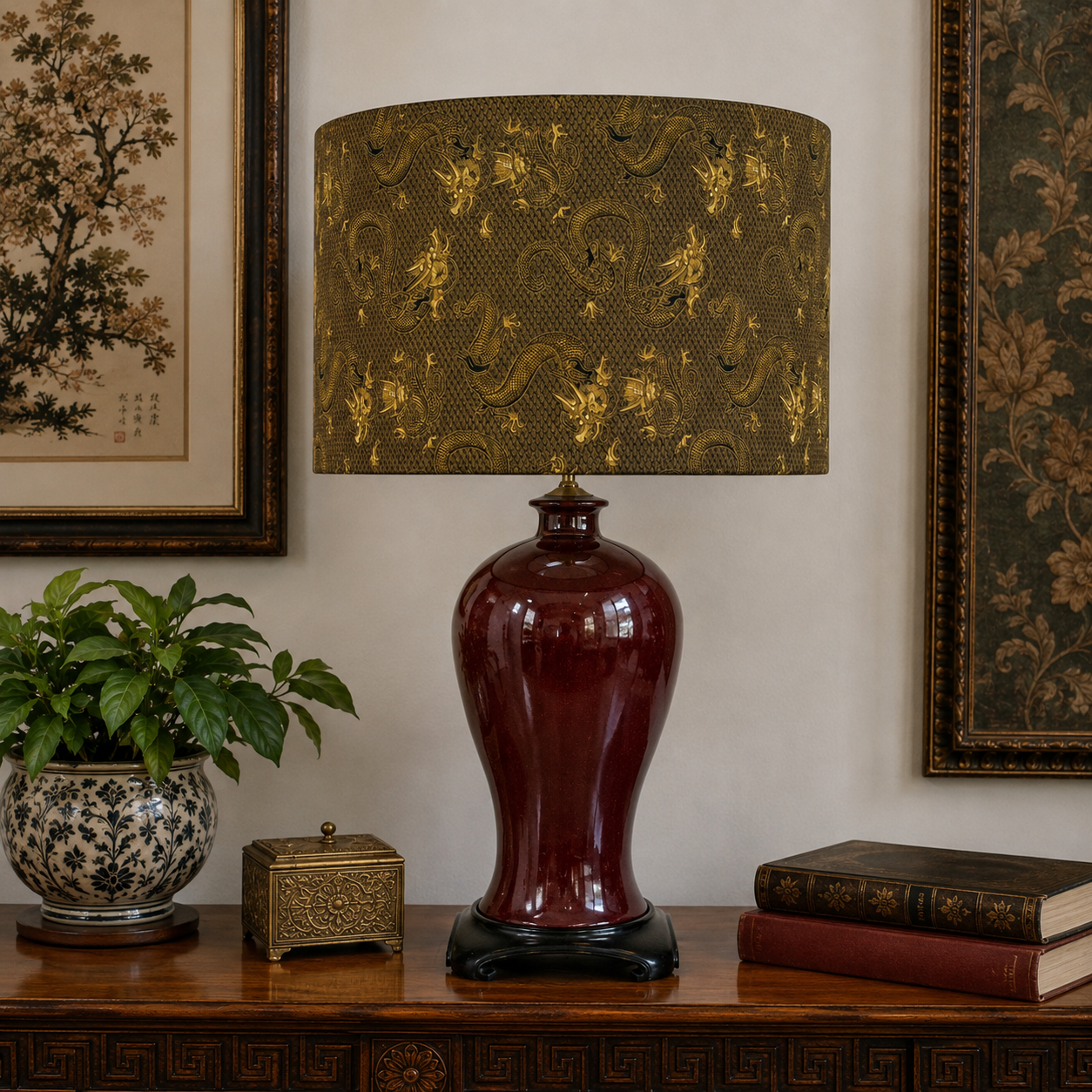 A table lamp with a glossy dark red ceramic base and a drum shade featuring a gold dragon pattern, styled on a wooden console table with a potted plant and books.