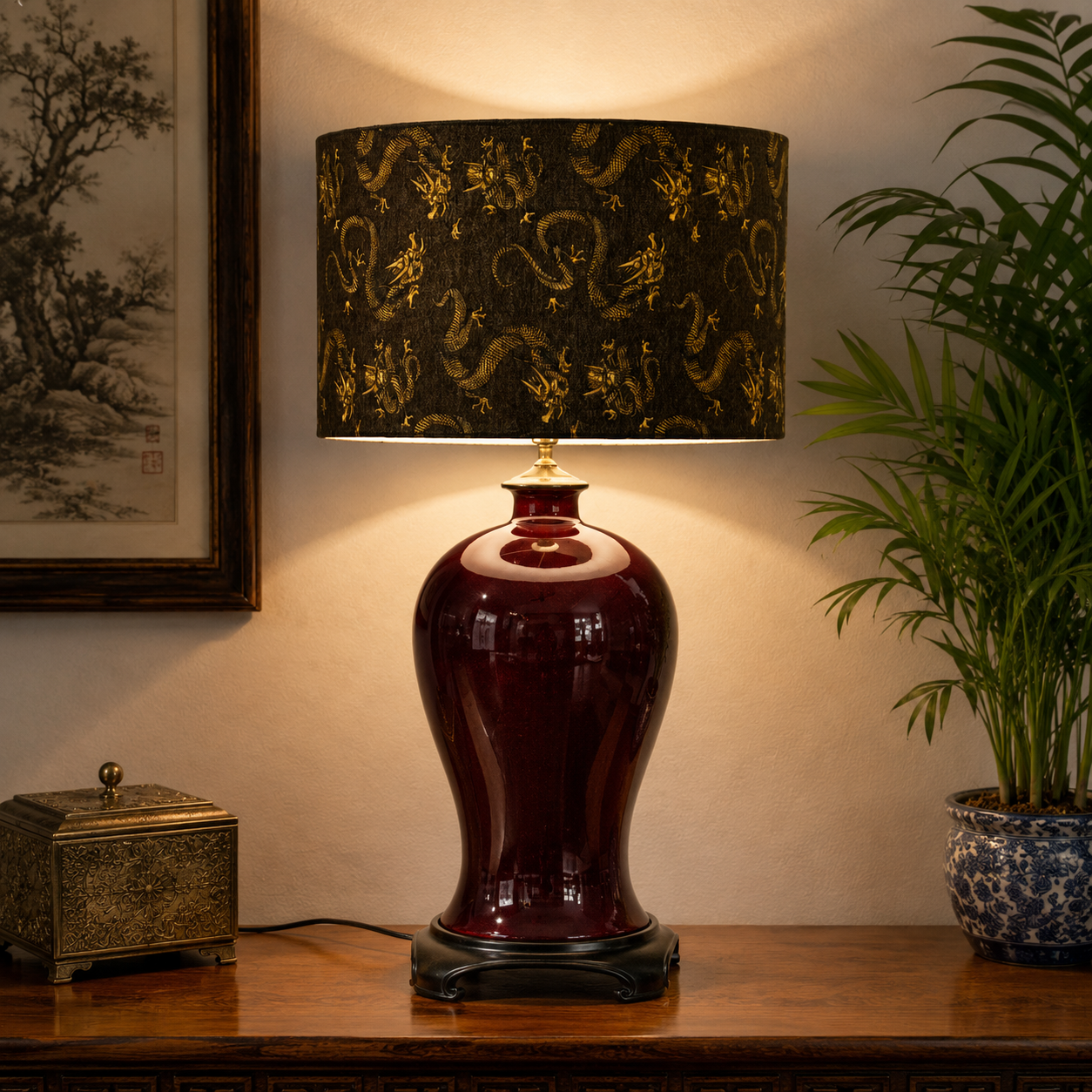 A table lamp with a glossy dark red ceramic base and a black drum shade decorated with gold dragon motifs, placed on a wooden console table in a styled room with a framed print and a potted plant.