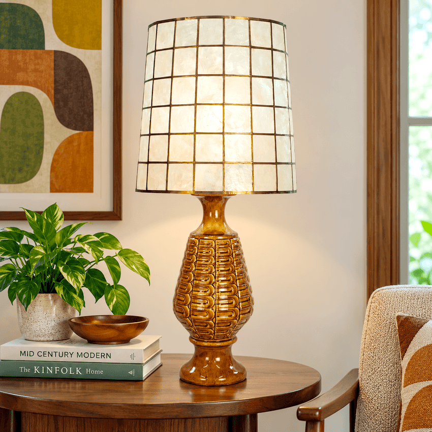 A table lamp with a textured amber-brown ceramic base and a square grid shade made of warm cream-colored glass panels, called capiz, shown lit on a wooden side table in a cozy room setting.