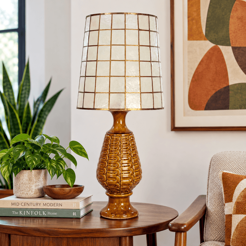 A table lamp with a textured amber-brown ceramic base and a cream-colored grid-patterned shade with gold-toned lines, styled on a wooden side table in a cozy living room setting.
