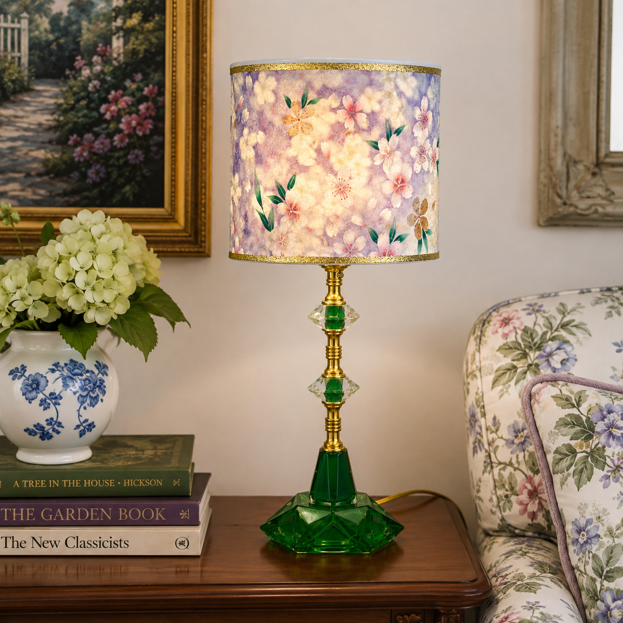 A decorative table lamp with a floral-patterned drum shade in pastel lavender, cream, and pink, featuring a gold-toned stem and a faceted green glass base, shown on a wooden side table in a living room setting.