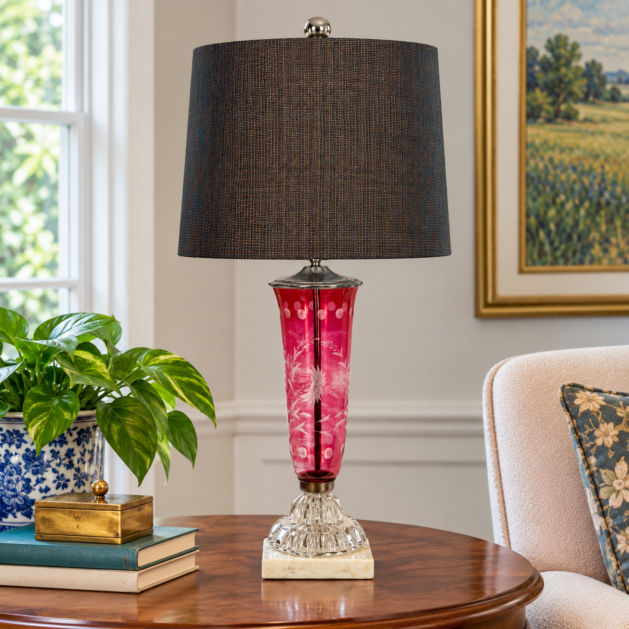 A table lamp with a dark charcoal gray fabric drum shade and a sculptural pink glass base with white floral details and a crystal pedestal, displayed on a wooden table in a living room setting.