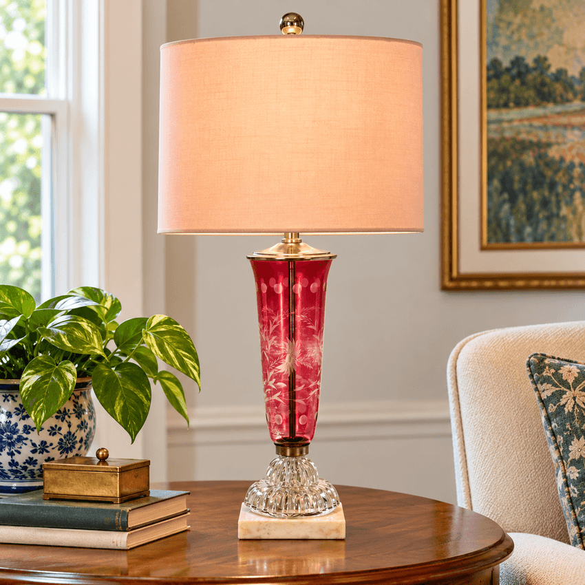 A table lamp with a warm peach fabric drum shade, a red patterned glass vase body, and a clear faceted crystal base on a round wooden table in a living room setting.