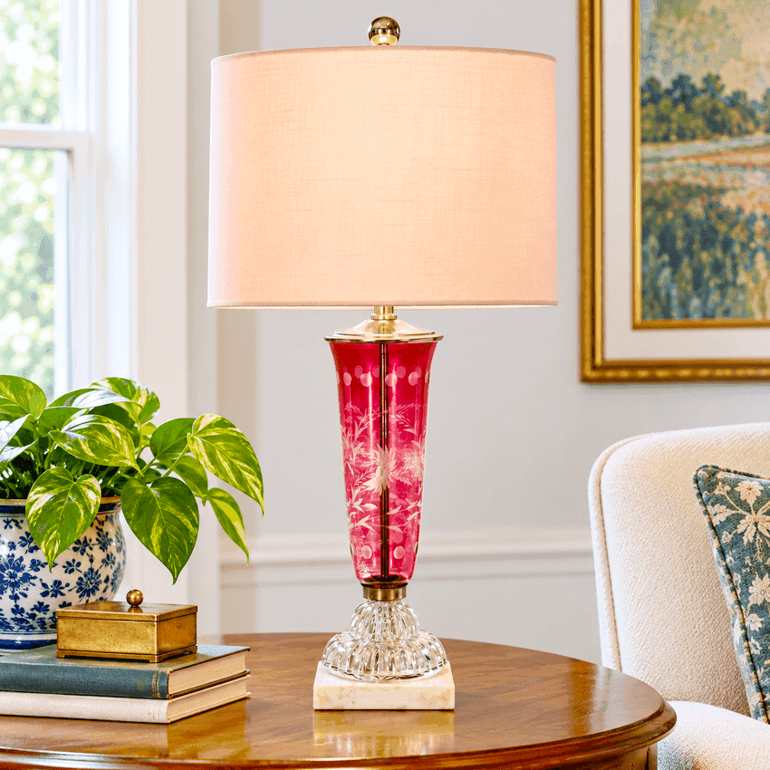 A table lamp with a warm peach fabric drum shade, a red patterned glass vase body, and a clear faceted crystal base on a round wooden table in a living room setting.