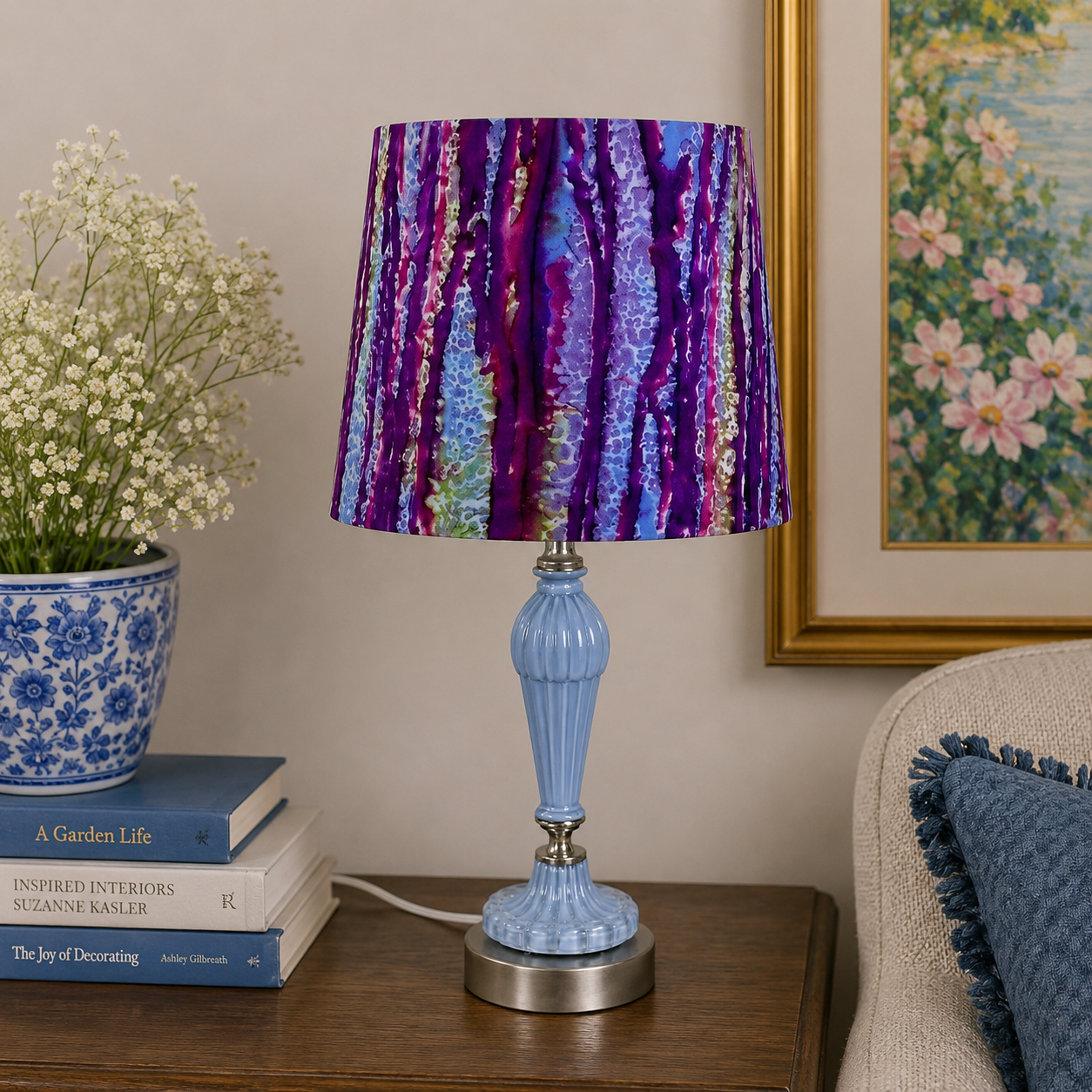A table lamp with a light blue fluted base and a pleated drum shade featuring a purple, blue, and gold abstract tree pattern, styled on a wooden side table in a living room setting.