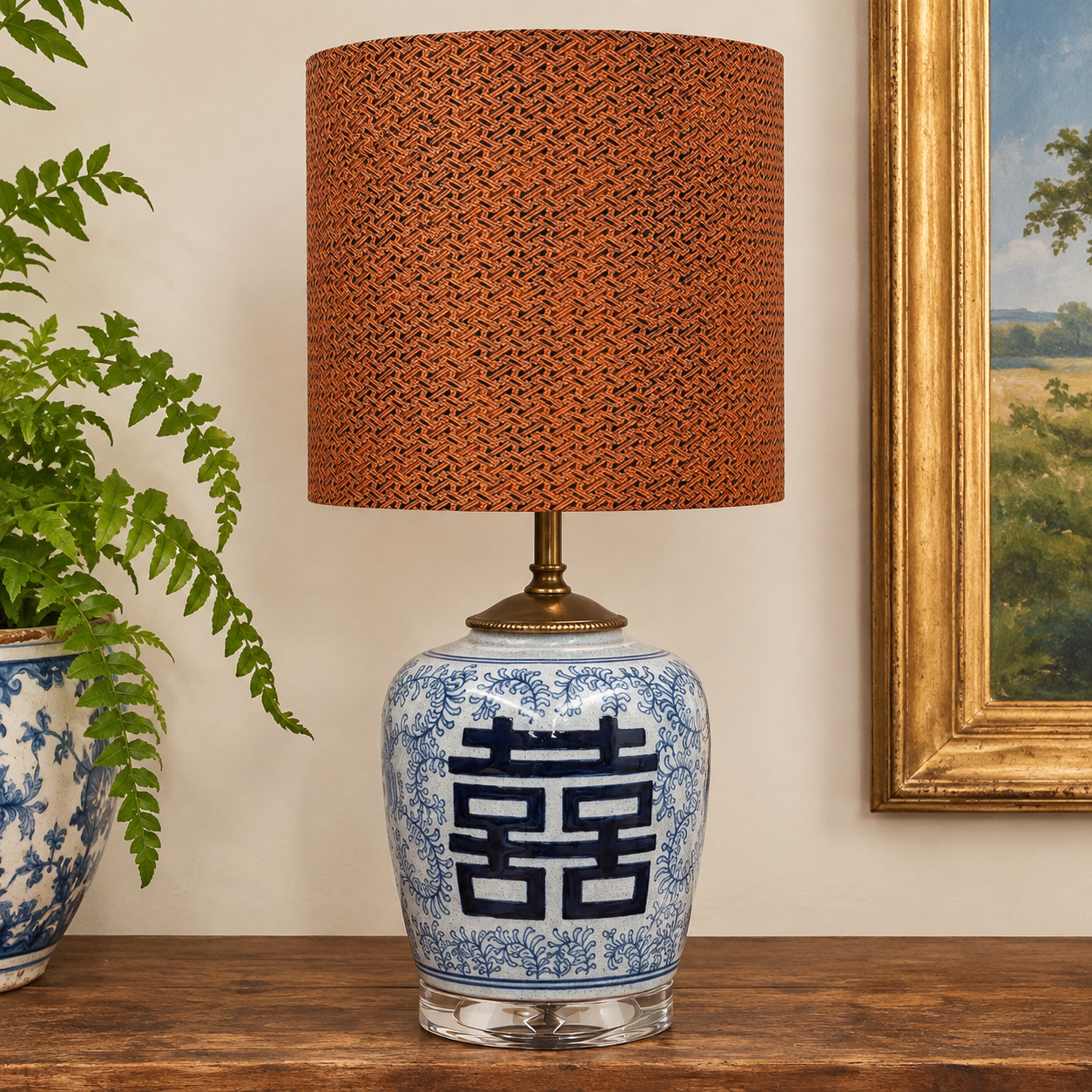 A table lamp with a blue and white porcelain-style ceramic base featuring a large dark blue double happiness symbol, topped with a textured rust-orange drum shade and a brass-toned neck, shown on a wooden surface against a beige wall with a plant and framed artwork nearby.