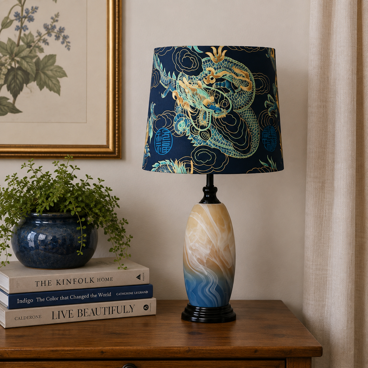 A table lamp with a navy blue drum shade featuring a detailed green and gold dragon design, and a marbled ceramic base in beige, cream, and blue, styled on a wooden side table next to a potted plant and books.
