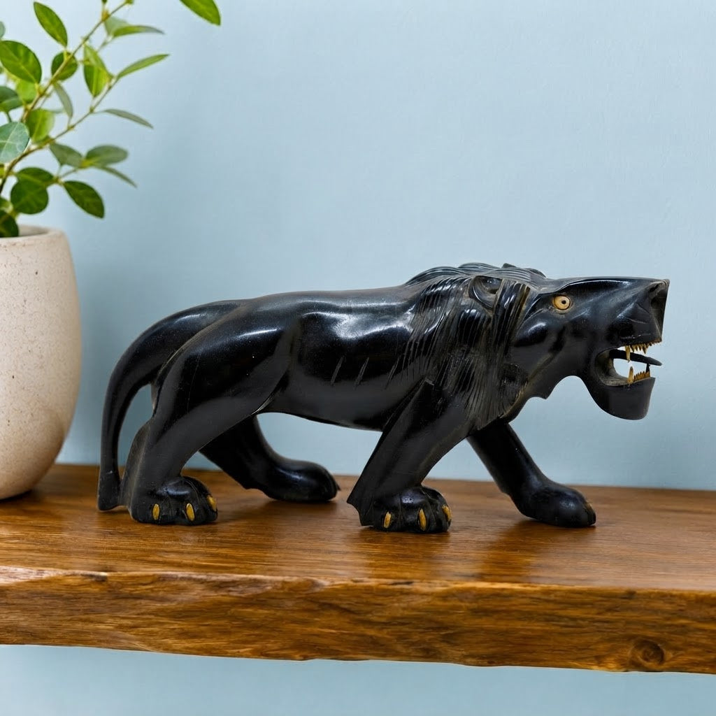 A carved black lion figurine with gold accents on its paws, teeth, and eyes, standing on a rustic wooden shelf next to a potted plant against a light blue wall.