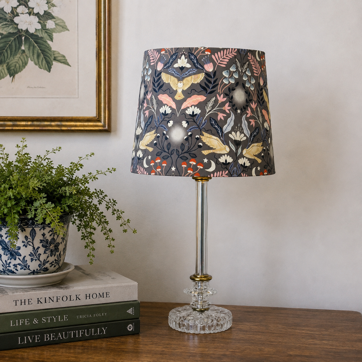 A table lamp with a dark floral and bird-patterned fabric shade, a clear glass stem, and a clear faceted glass base, styled on a wooden table next to a potted plant and books.