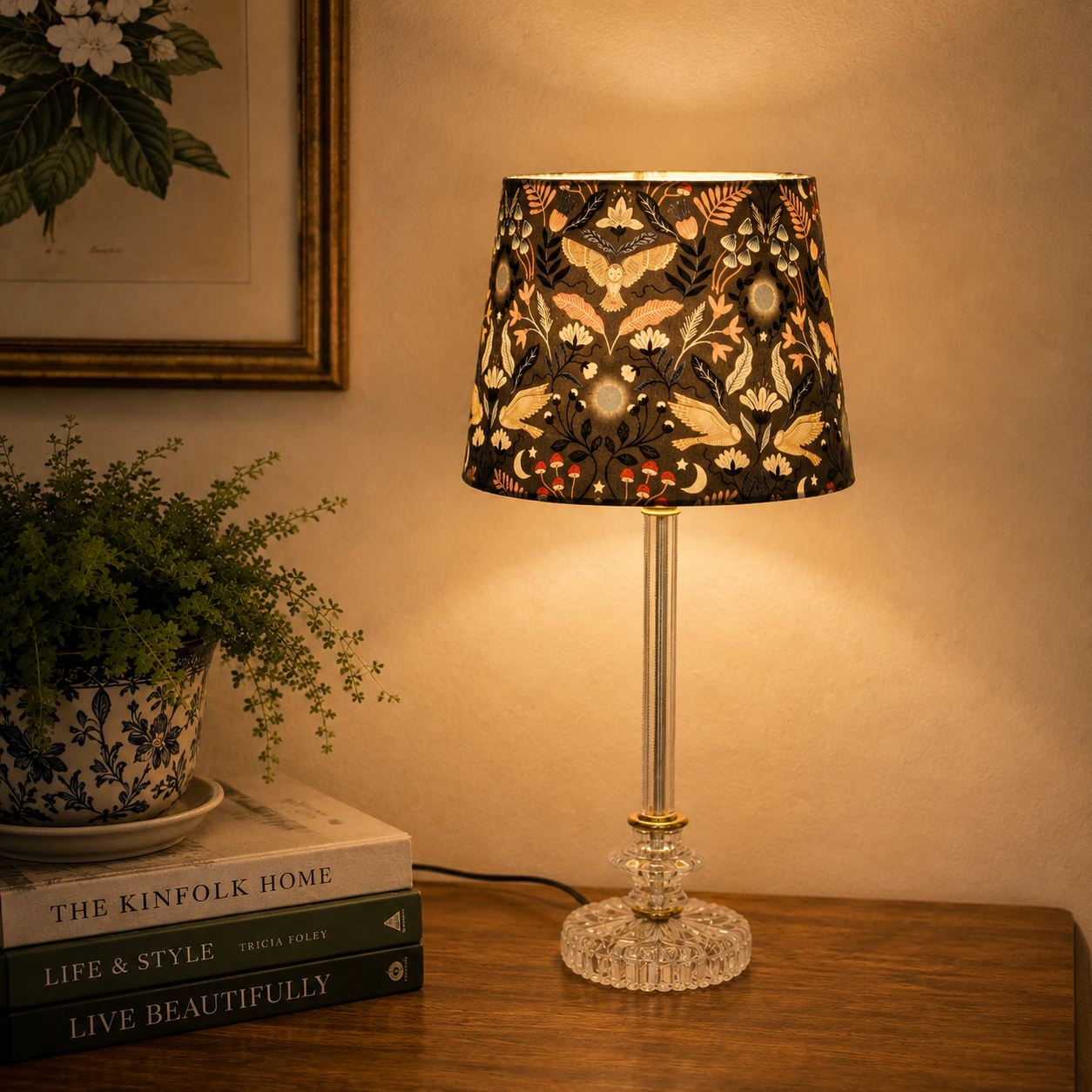 A table lamp with a black fabric shade featuring a warm-toned floral and butterfly cutout pattern, set on a wooden table next to a potted plant and books in a cozy room.
