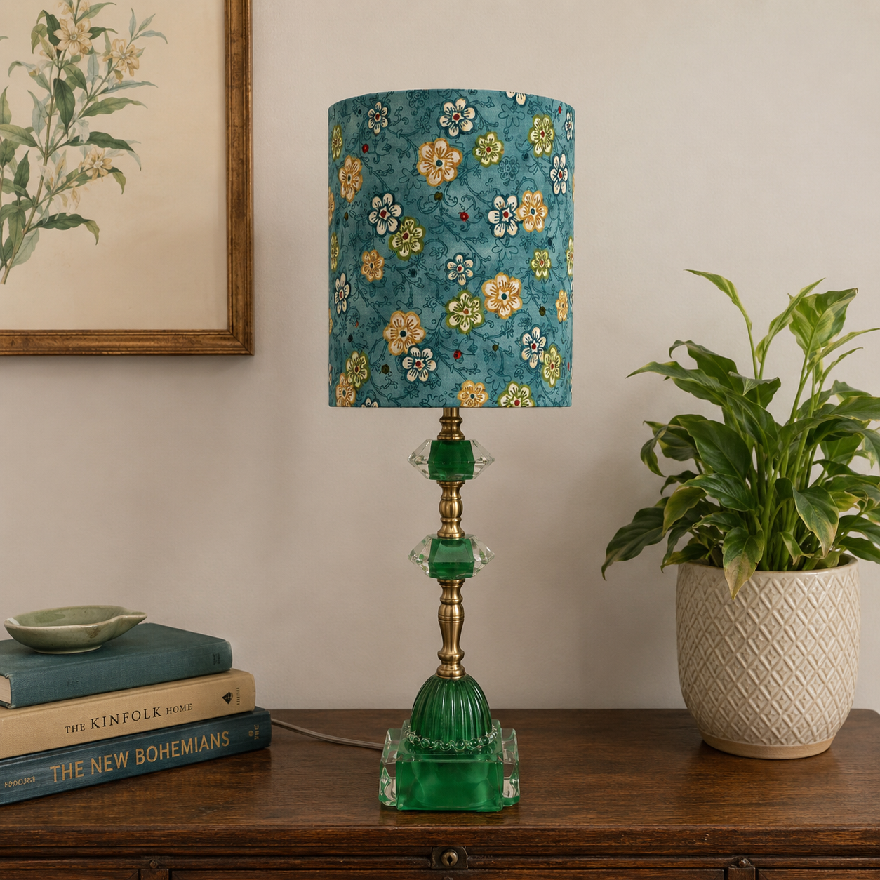 A table lamp with a teal floral drum shade and a green glass base sits on a wooden console table in a styled room setting, flanked by a potted plant and books.