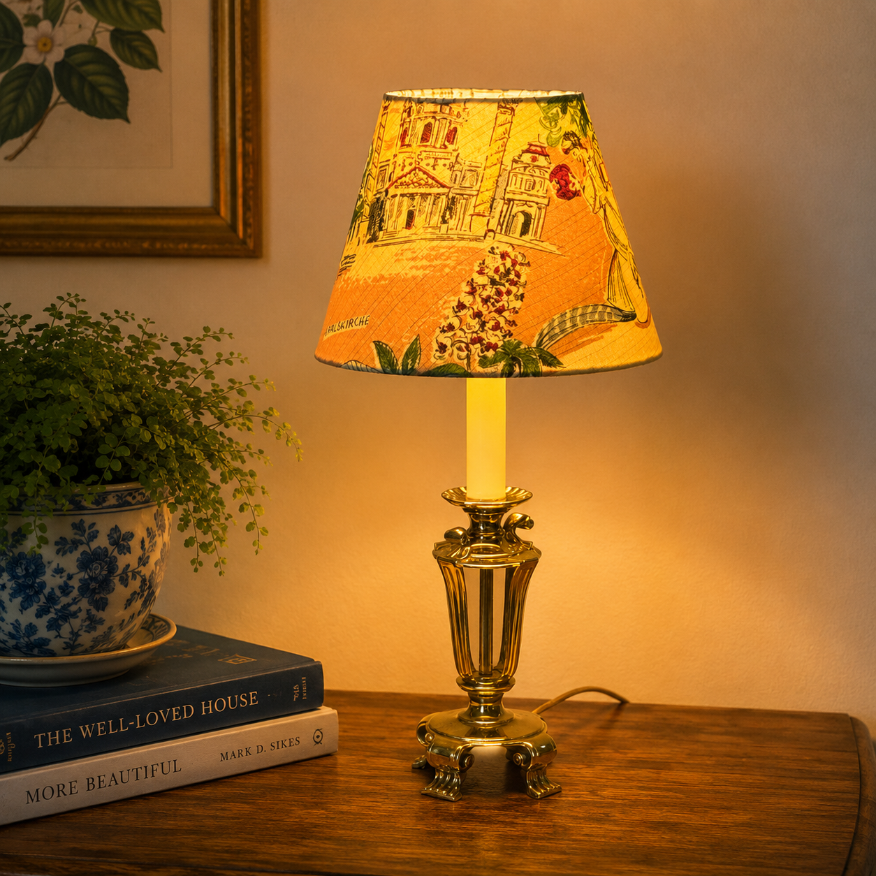 A table lamp with a warm yellow glowing bulb and a patterned lampshade featuring a classical building and Christmas tree, set on a wooden table next to a potted plant and books.