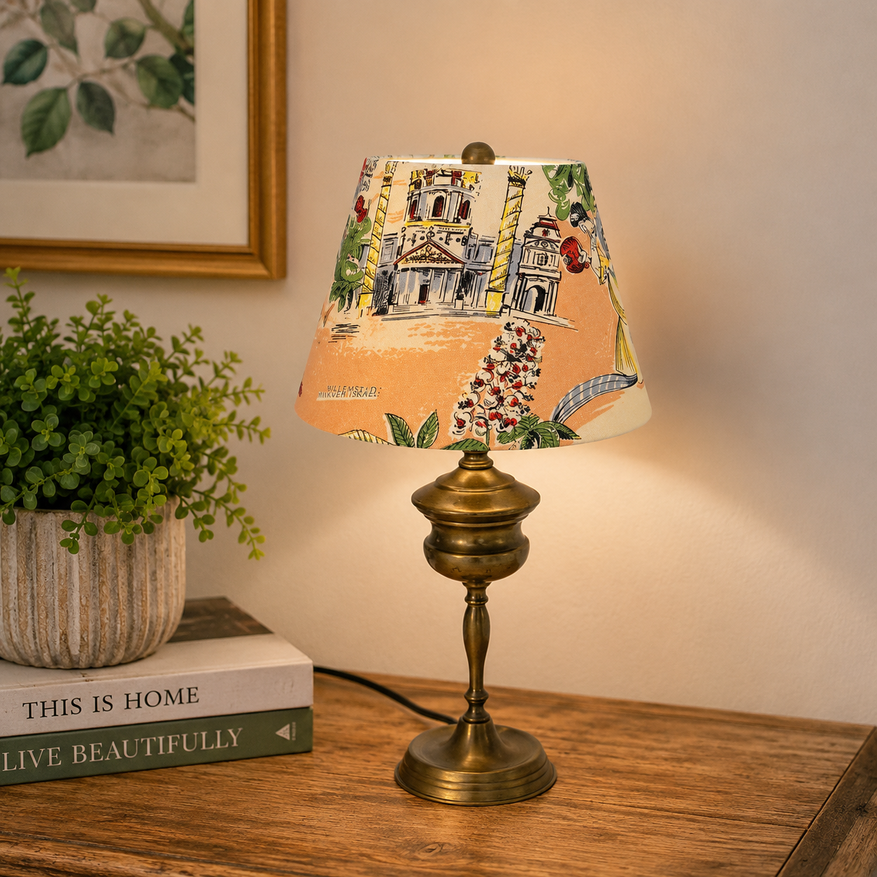 A table lamp with a brass-finished base and a conical lampshade printed with a colorful architectural scene, shown lit on a wooden table beside a potted plant and books against a warm beige wall.