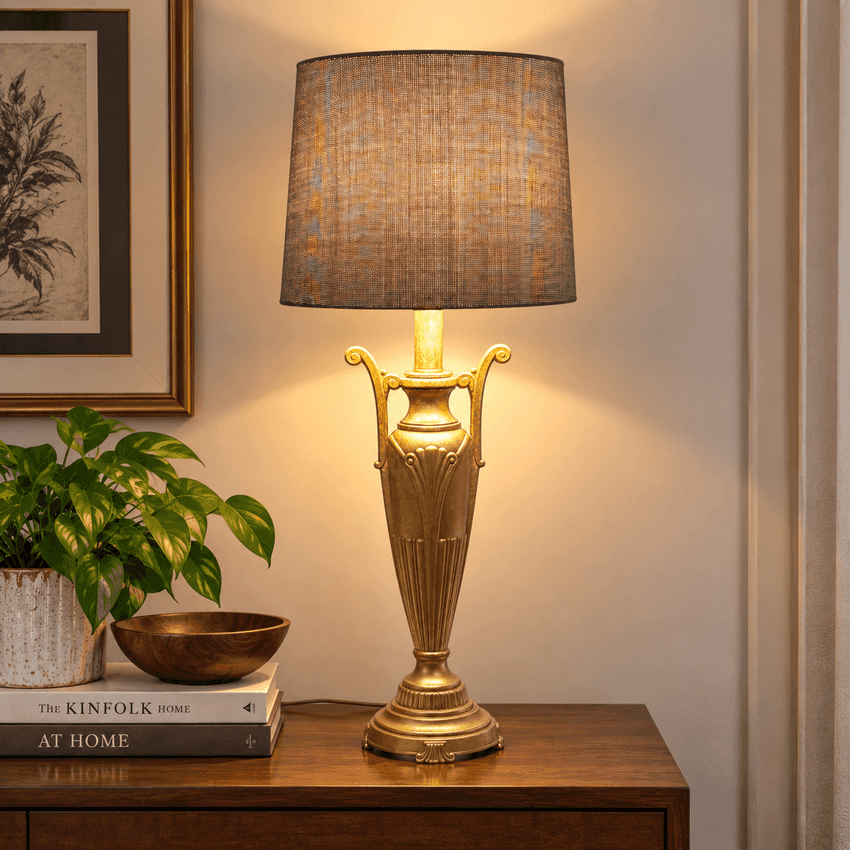 A classic table lamp with a gold-finished urn-style base and a textured brown fabric drum shade, illuminated and placed on a wooden side table in a styled room setting.