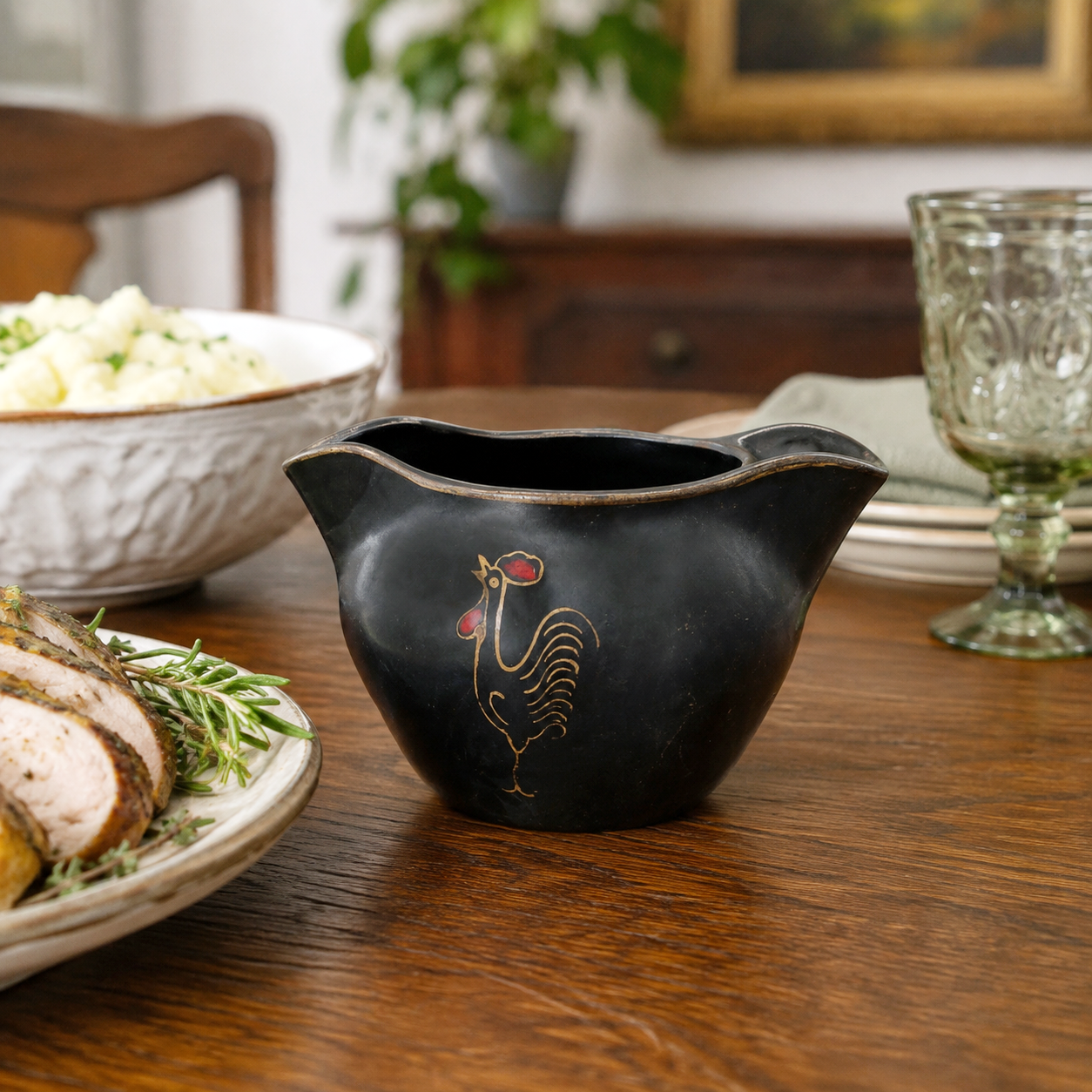 A black ceramic gravy boat with a gold rooster design and red accents, set on a wooden dining table with food and tableware in the background.