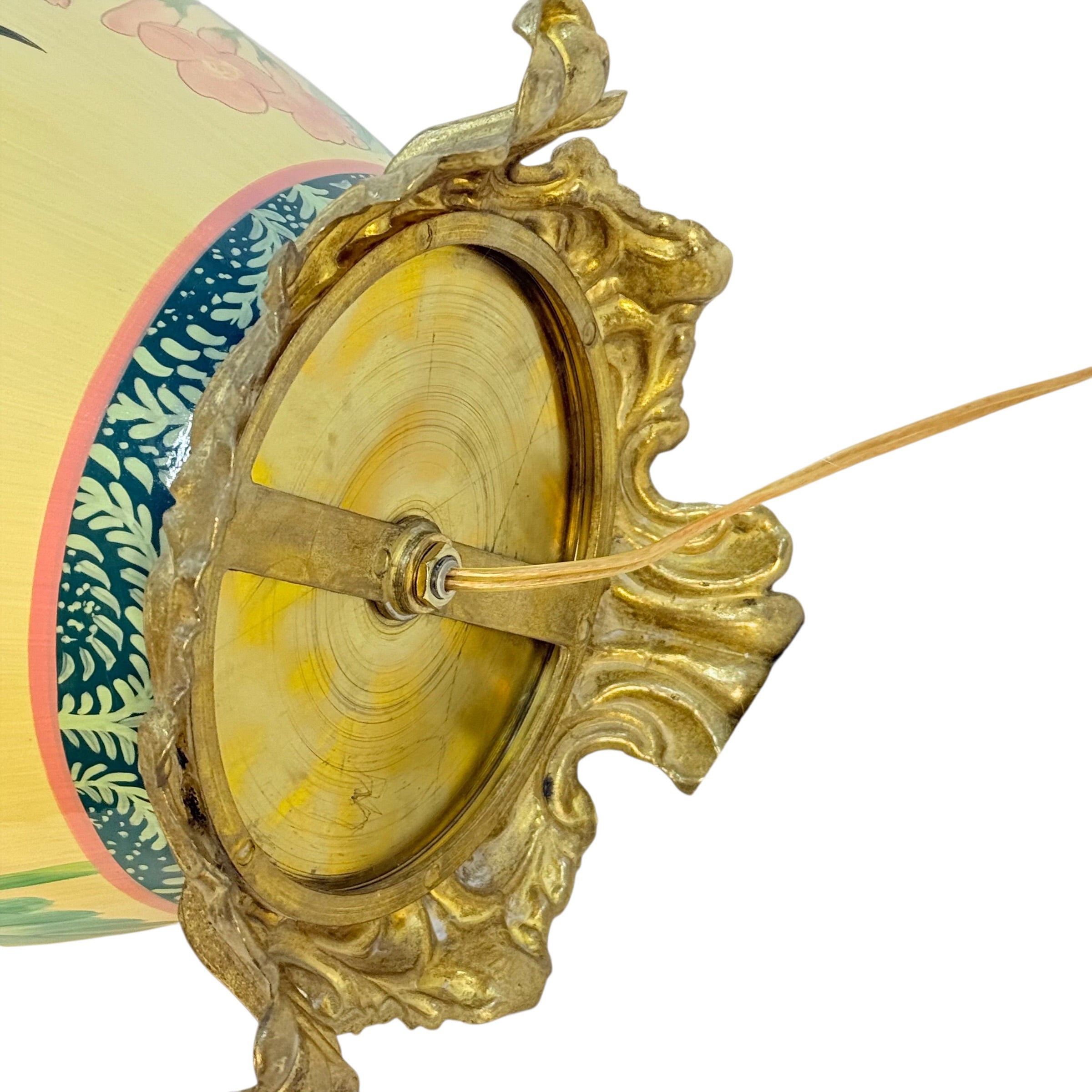Close-up detail of a decorative hanging lantern or drum-like ornament with an ornate gold metal frame, a round yellow center, and part of a painted body showing blue, green, and pink floral patterns on a white background.