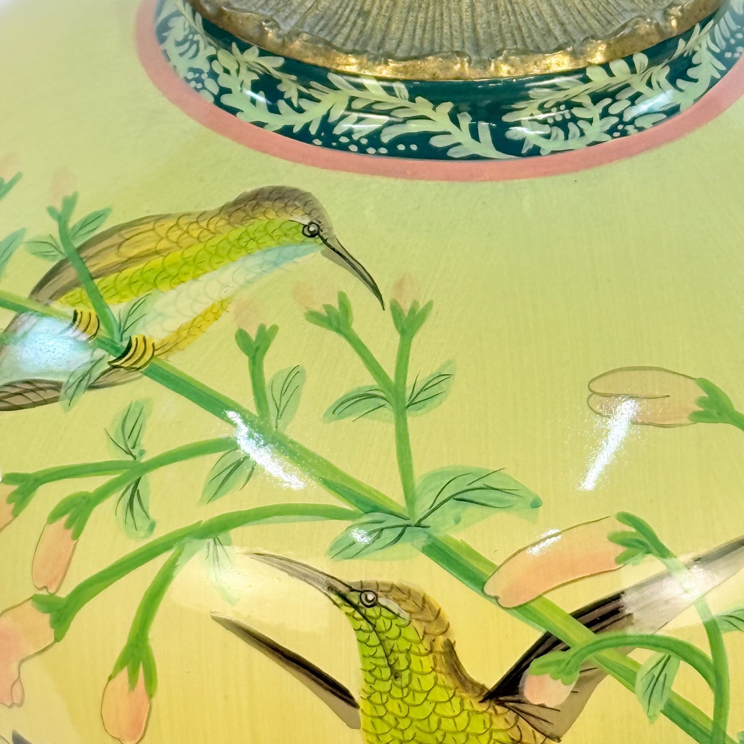 Close-up of a decorative painted surface featuring two green and yellow birds perched on flowering branches, with a pale yellow ground and part of a blue-and-gold circular motif visible at the top.