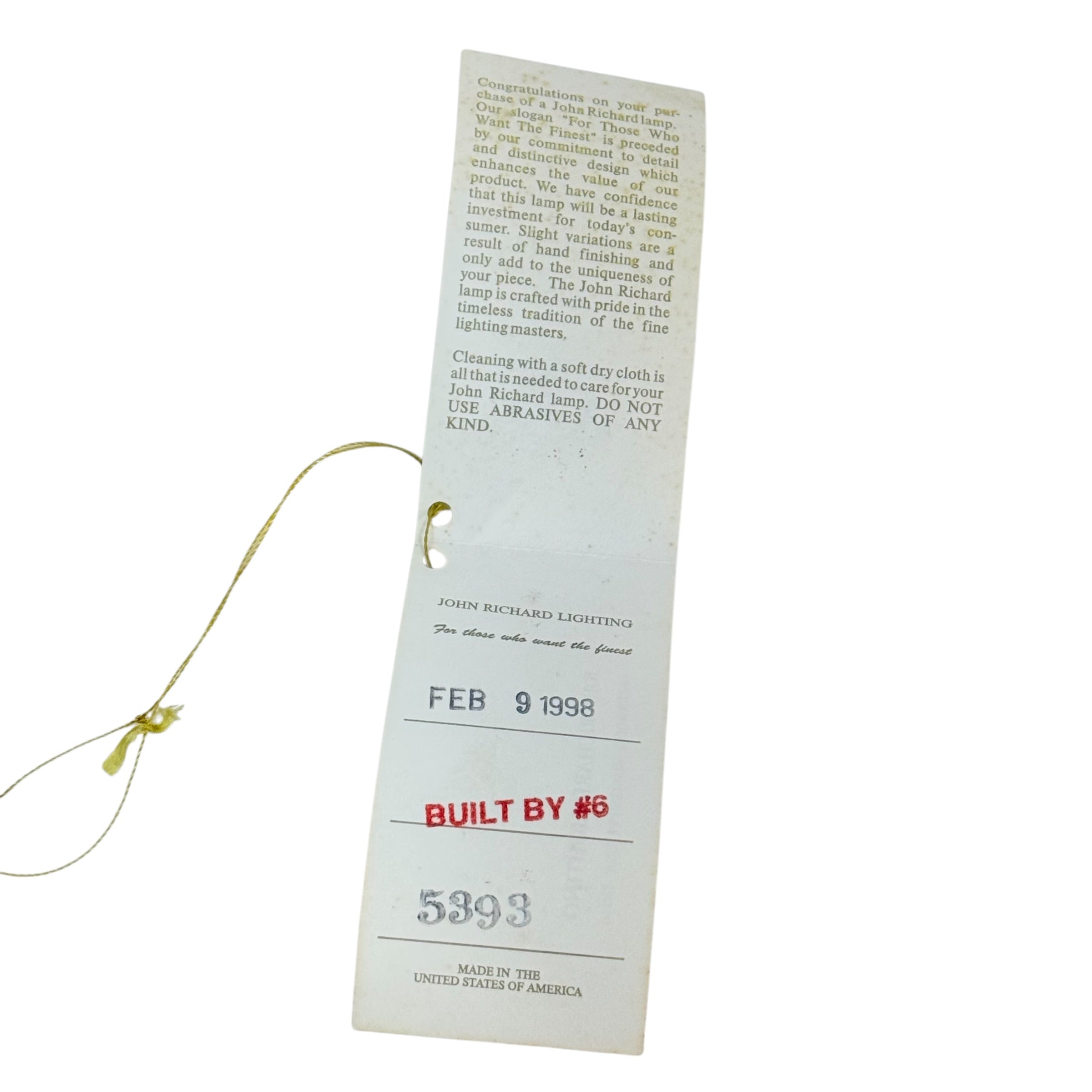 A vintage paper hangtag from John Richard Lighting Company, dated February 9, 1998, with printed text and a red 'BUILT BY' stamp, attached to a gold-colored string on a white background.