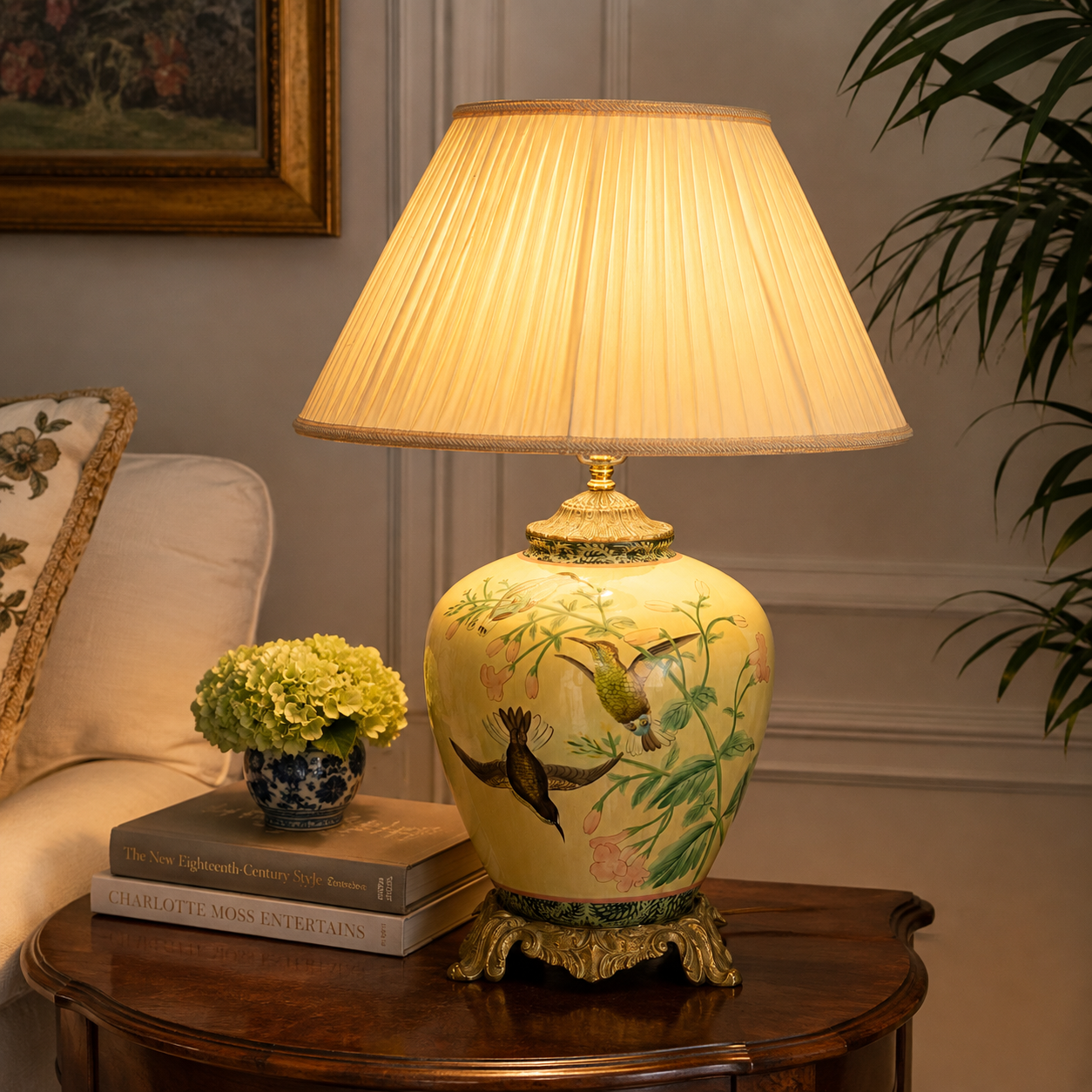 A decorative table lamp with a pleated beige shade and a yellow ceramic base featuring painted birds and foliage, placed on a wooden side table in a living room setting.