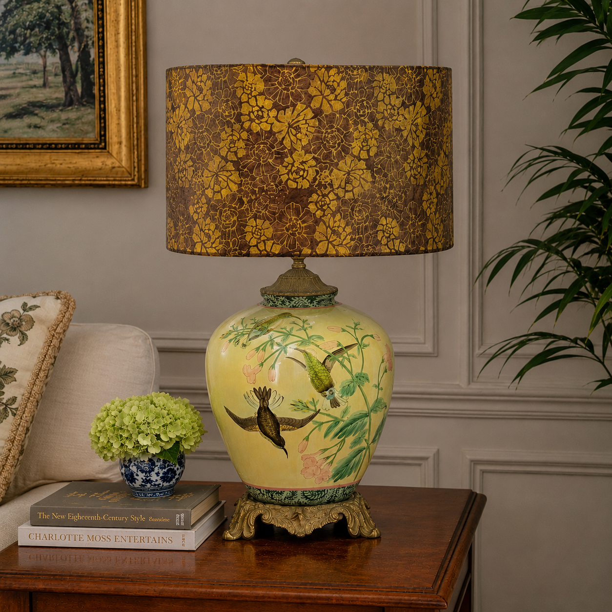 An ornate table lamp with a yellow-green ceramic body decorated with birds and floral motifs, topped with a brown and gold floral patterned drum shade, displayed on a wooden side table in a styled room setting.
