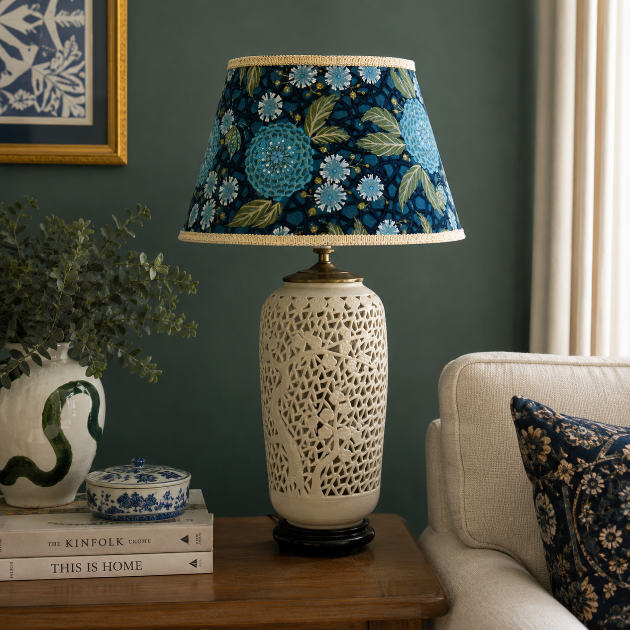 A decorative table lamp with a cream-colored carved ceramic base and a blue floral patterned fabric shade, styled on a wooden side table in a living room setting.
