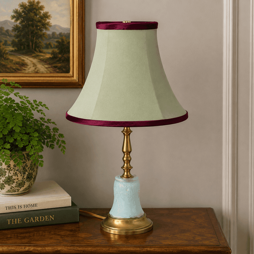 A table lamp with a pale sage green fabric shade trimmed in deep burgundy, a gold-toned metal stem, and a light blue marbled base, displayed on a wooden table in a styled room setting.