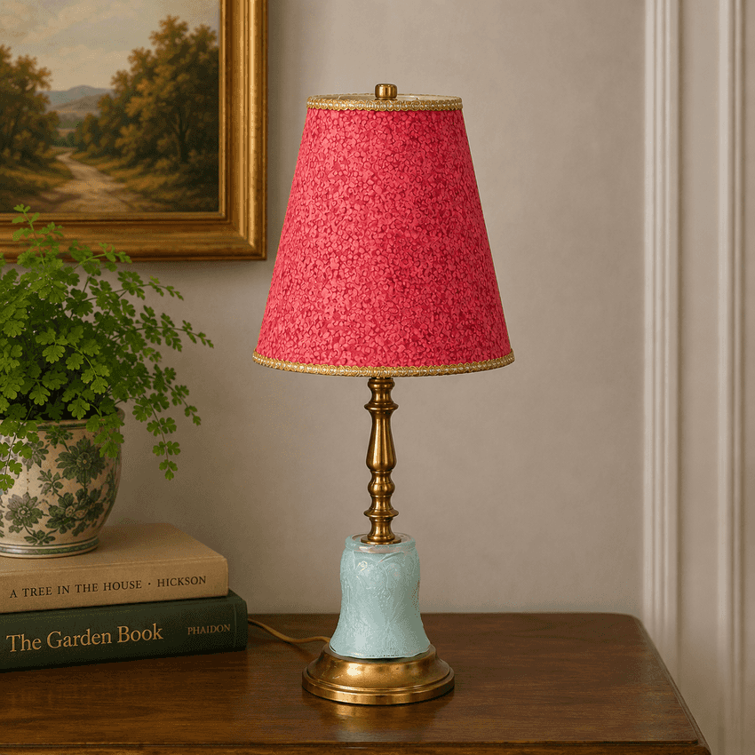 A table lamp with a textured pink shade, a brass-toned stem, and a light blue ceramic base, styled on a wooden table next to a potted plant and books.