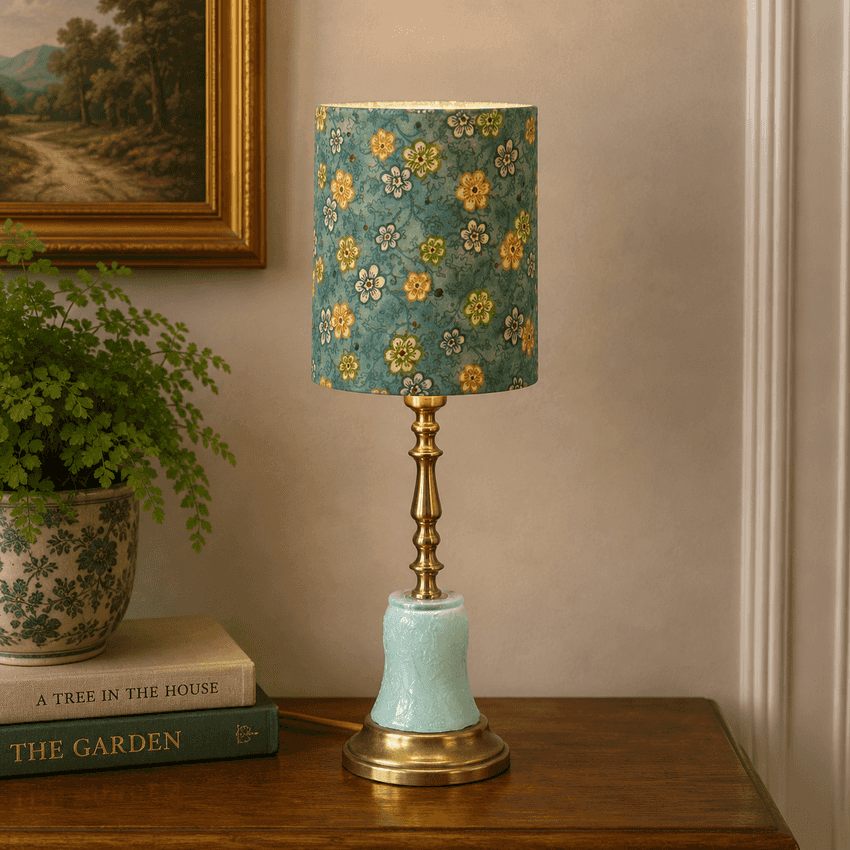 A vintage-style table lamp with a teal floral drum shade, a brass-toned stem, and a pale aqua ceramic base, displayed on a wooden table in a styled room setting.