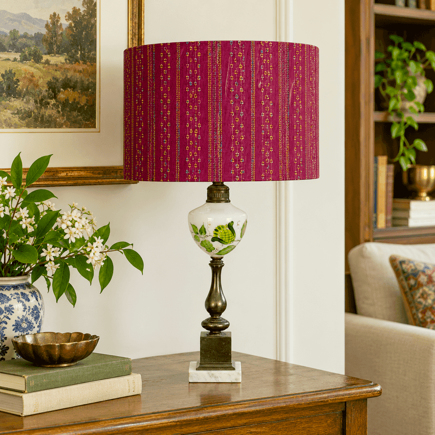 A table lamp with a deep red patterned fabric shade, a white ceramic body with green floral motifs, and a dark metal pedestal base, styled on a wooden console table in a living room setting.