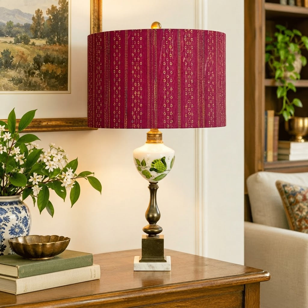 A lit table lamp with a deep red patterned fabric shade, a white ceramic body with green floral motifs, and a dark metal pedestal base, styled on a wooden console table in a living room setting.