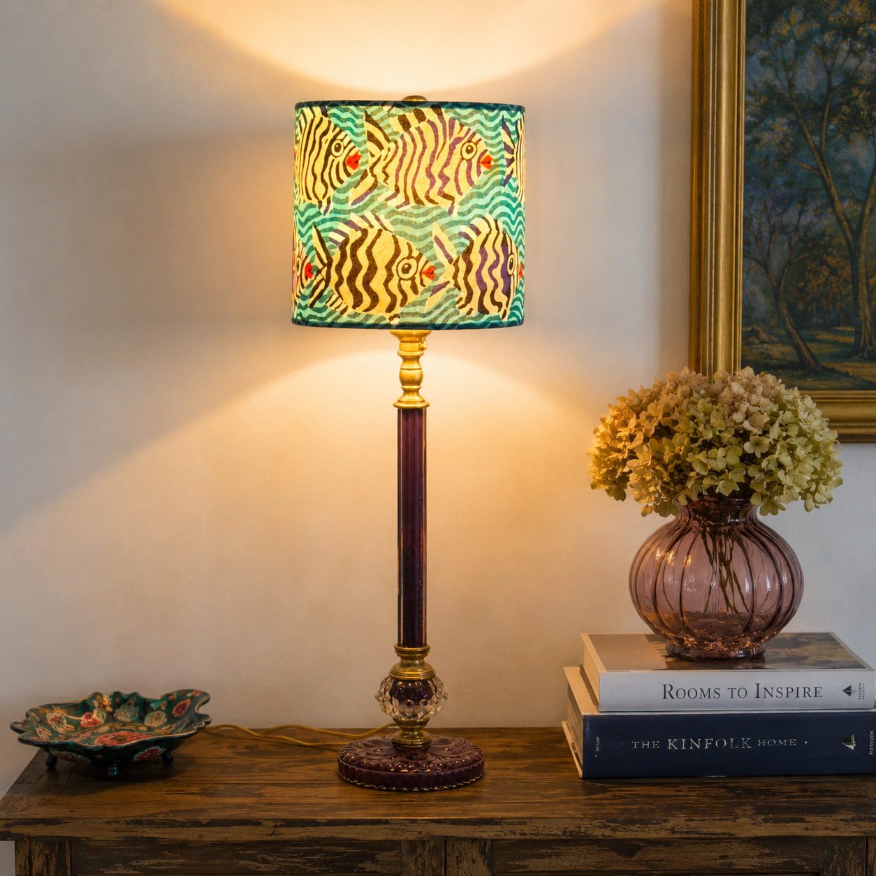 A decorative table lamp with a colorful fish-patterned drum shade and a dark purple column with gold accents, lit and placed on a wooden console table next to a pink glass vase with flowers.