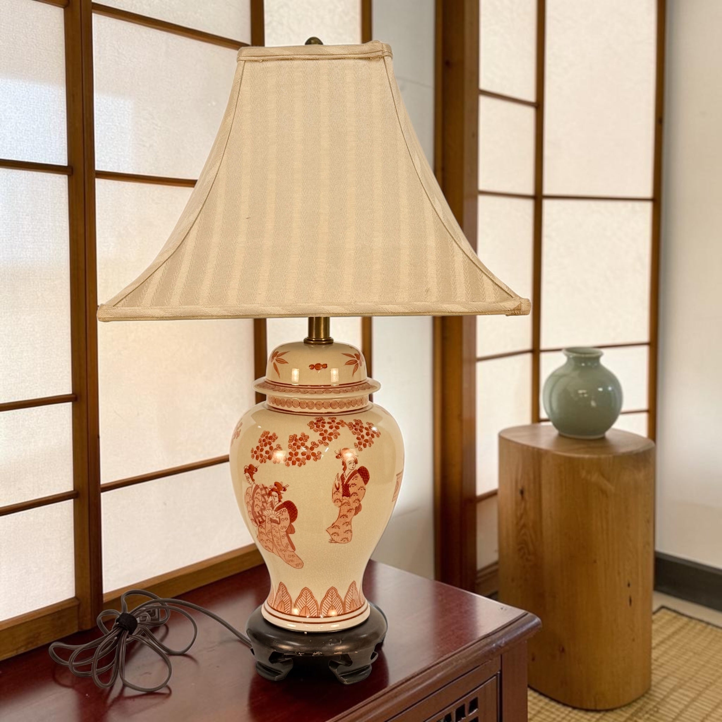 A decorative table lamp with a cream striped fabric shade and a glossy cream ceramic base decorated with red-orange figures and floral motifs, set on a dark wooden table in front of shoji screens.