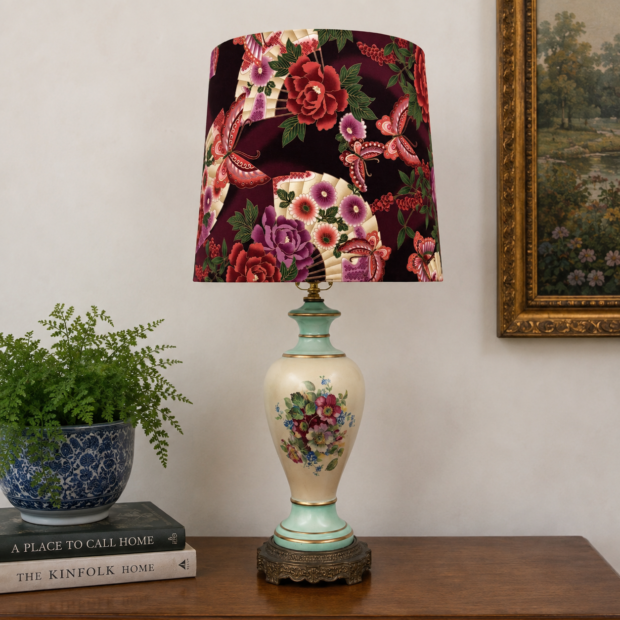 A decorative table lamp with a floral-patterned drum shade and a porcelain-style vase base, styled on a wooden table against a beige wall with a framed painting and a potted plant.